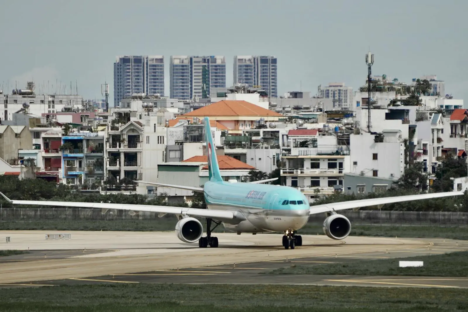 Photo by Pham Huynh Tuan Vy Airplane taxiing on runway at urban airport with cityscape in background.