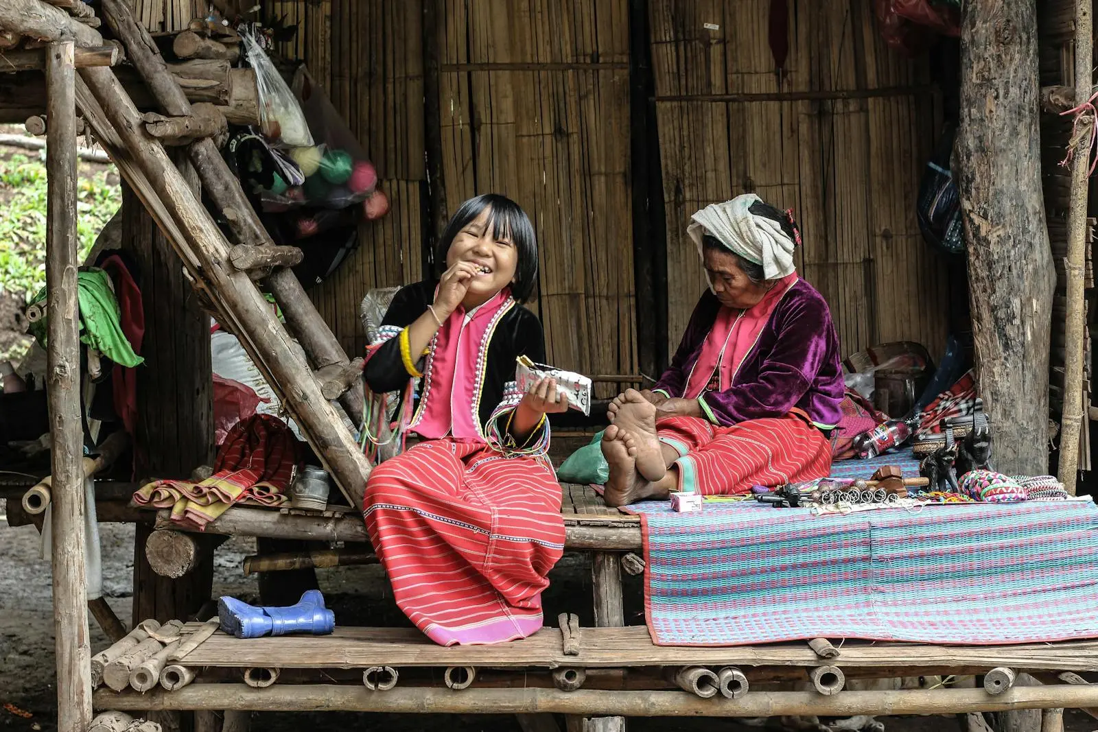 Asian girl smiling and elderly woman in traditional attire sitting outside a bamboo hut.