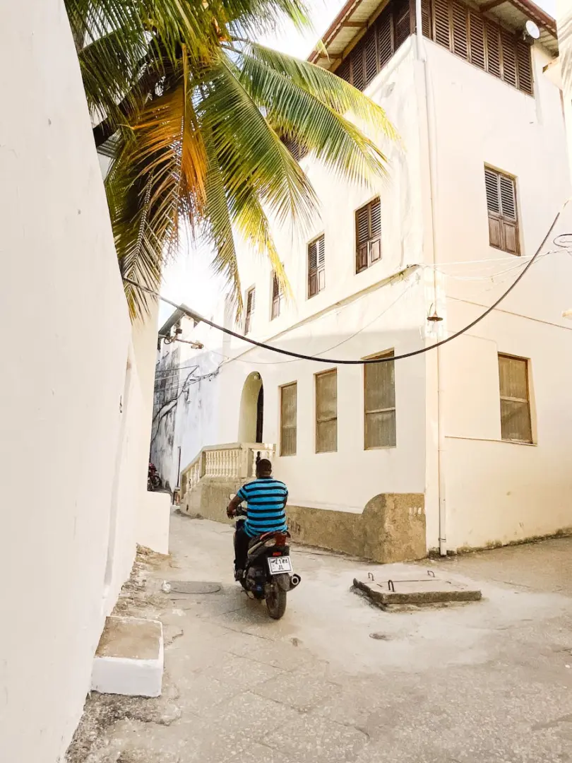 A man on a scooter in a narrow alleyway lined with tropical palm trees and buildings.