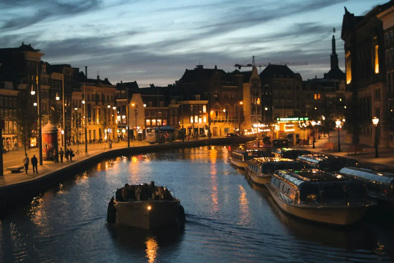 Photo by Artem Zhyzhyn Canal boats pass buildings illuminated at dusk.