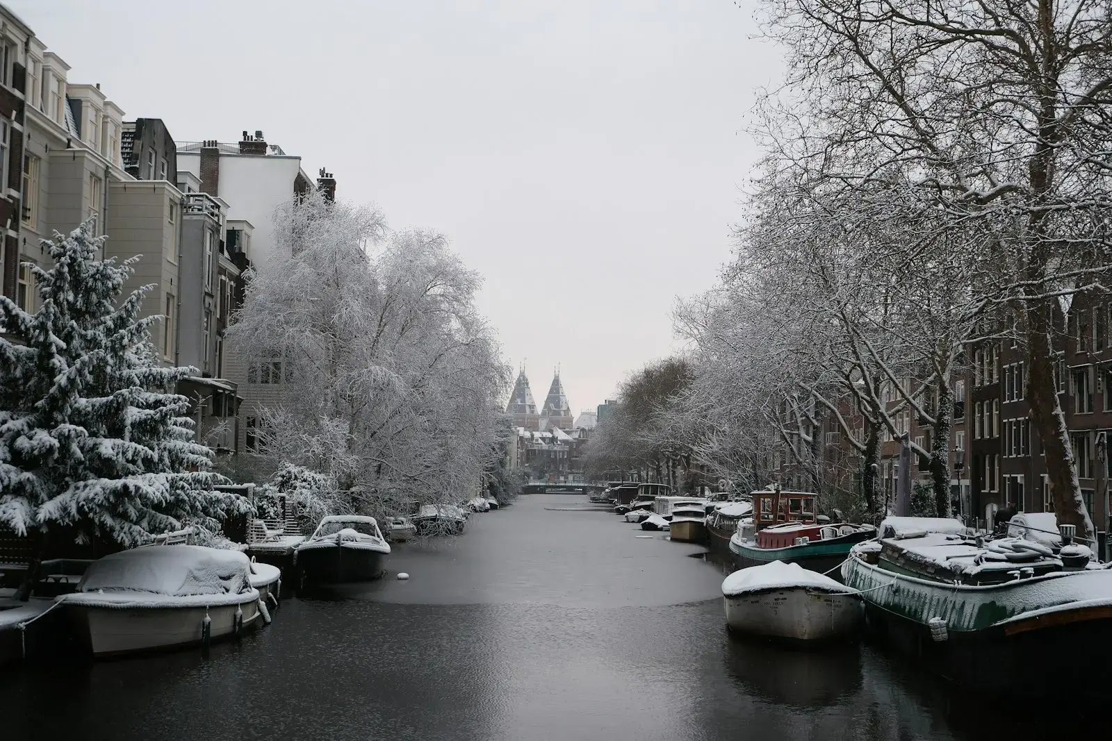 Photo by Farah Almazouni Snowy canal with boats and bare trees in amsterdam.