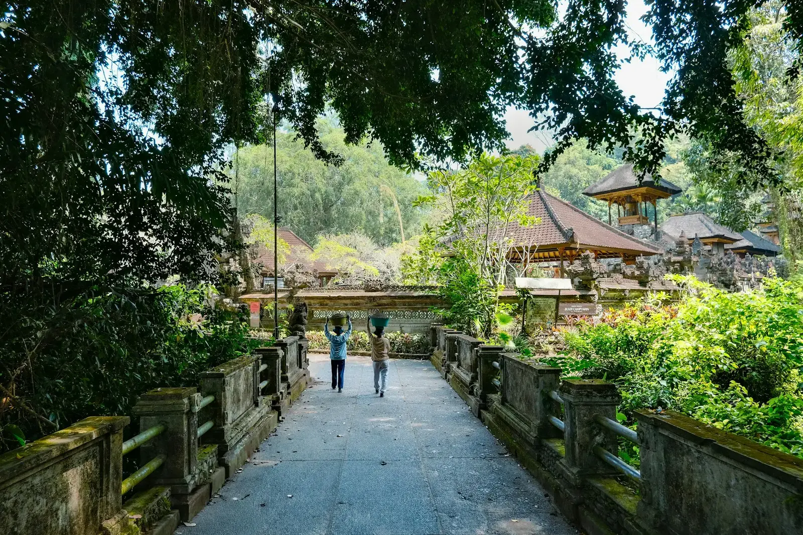 Two people walk across a stone bridge in lush greenery.