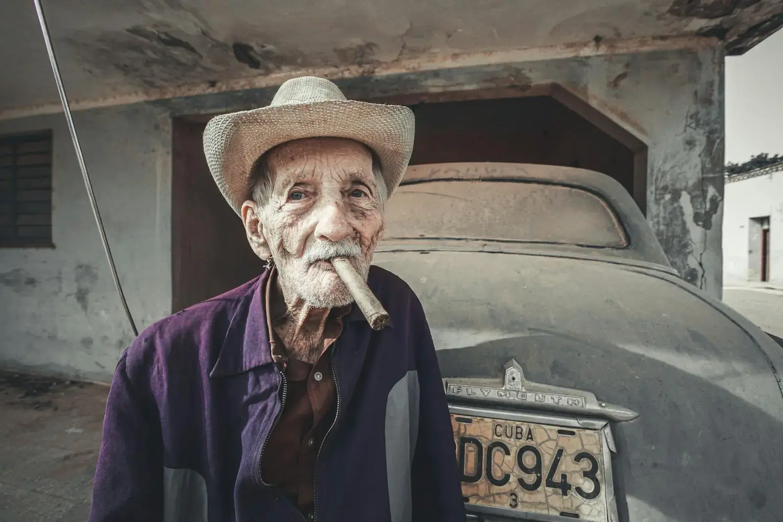 Portrait of an elderly man wearing a hat, smoking a cigar in front of a vintage car in Cuba.