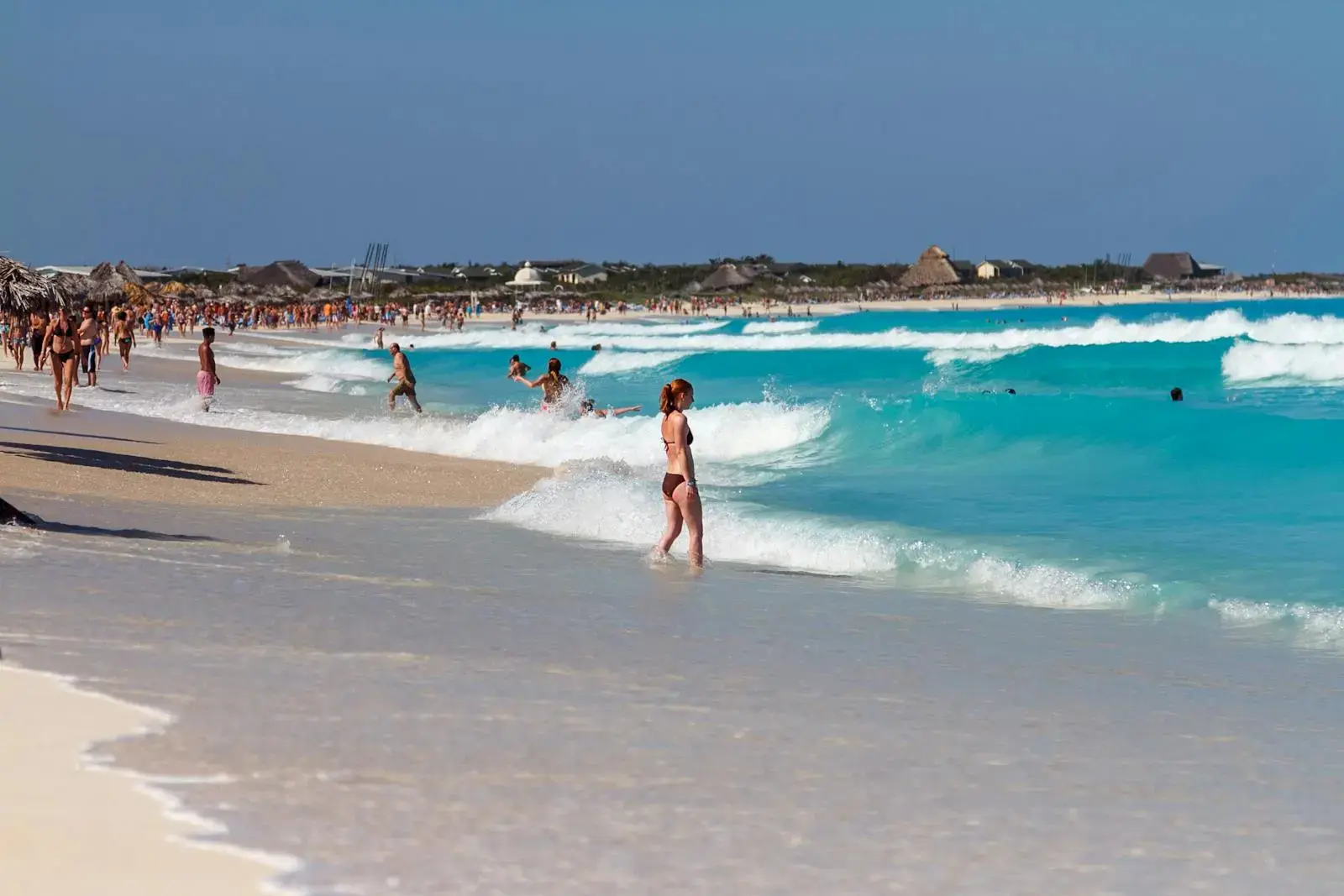 Crowded beach scene with turquoise ocean waves and people enjoying summer activities.