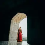 A woman in a traditional saree stands in a grand, ornate doorway with intricate patterns.