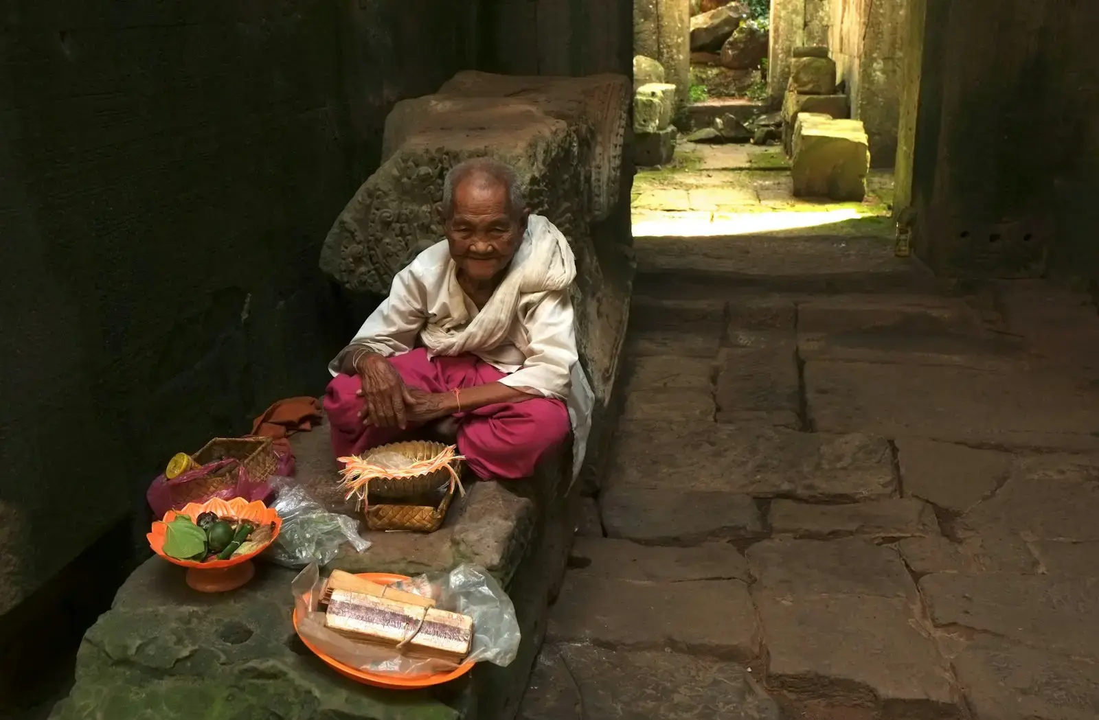 Photo by Boudewijn Huysmans Elderly person sitting with offerings in ancient ruins.