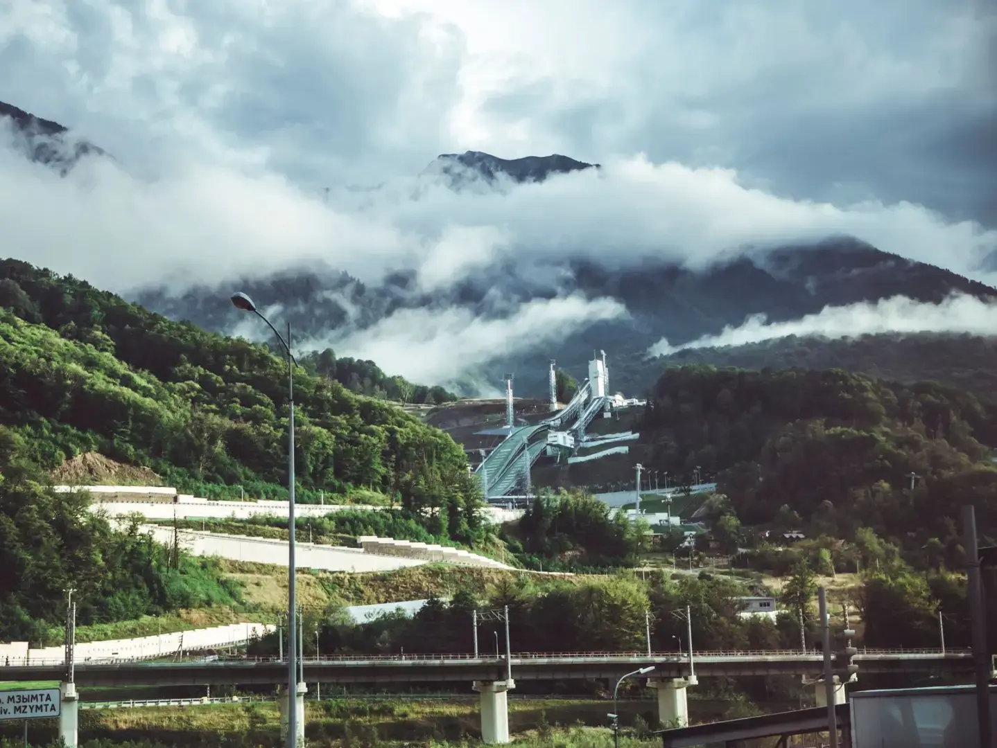 Photo by Mike Scenic mountain landscape featuring a bridge, lush greenery, and misty clouds covering peaks.
