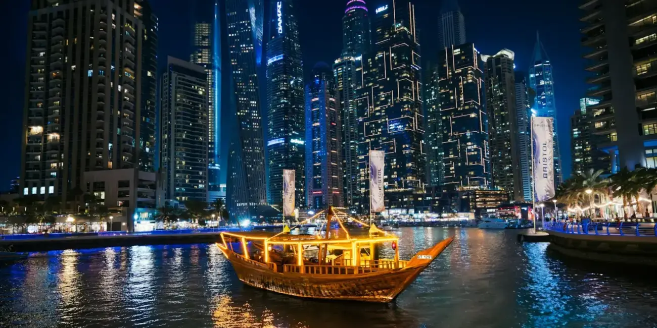 A traditional dhow boat sails past illuminated skyscrapers at night.