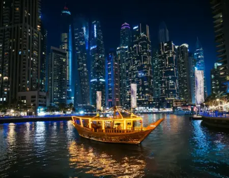 A traditional dhow boat sails past illuminated skyscrapers at night.