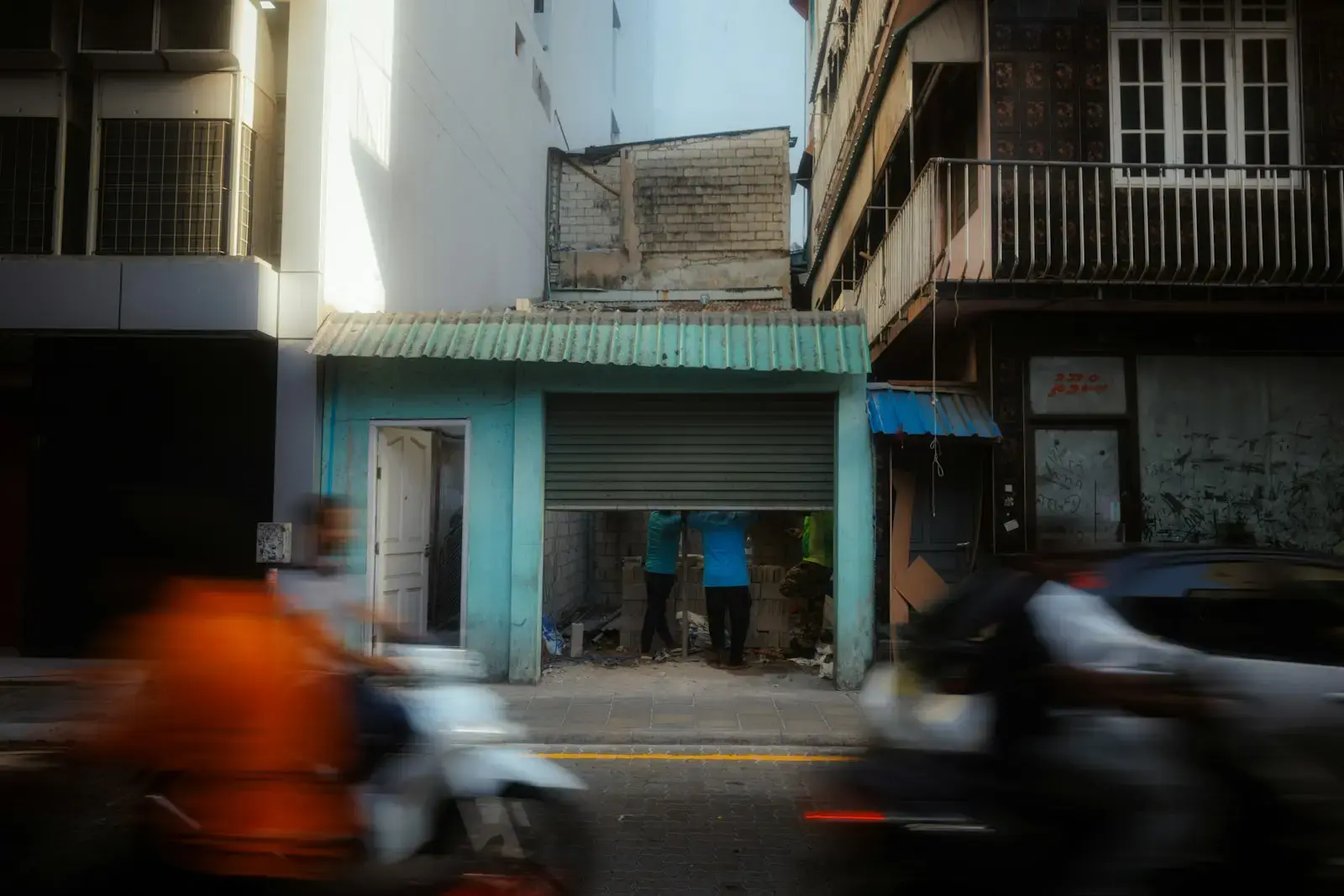 Motorcyclists pass a small shop with closed metal shutters