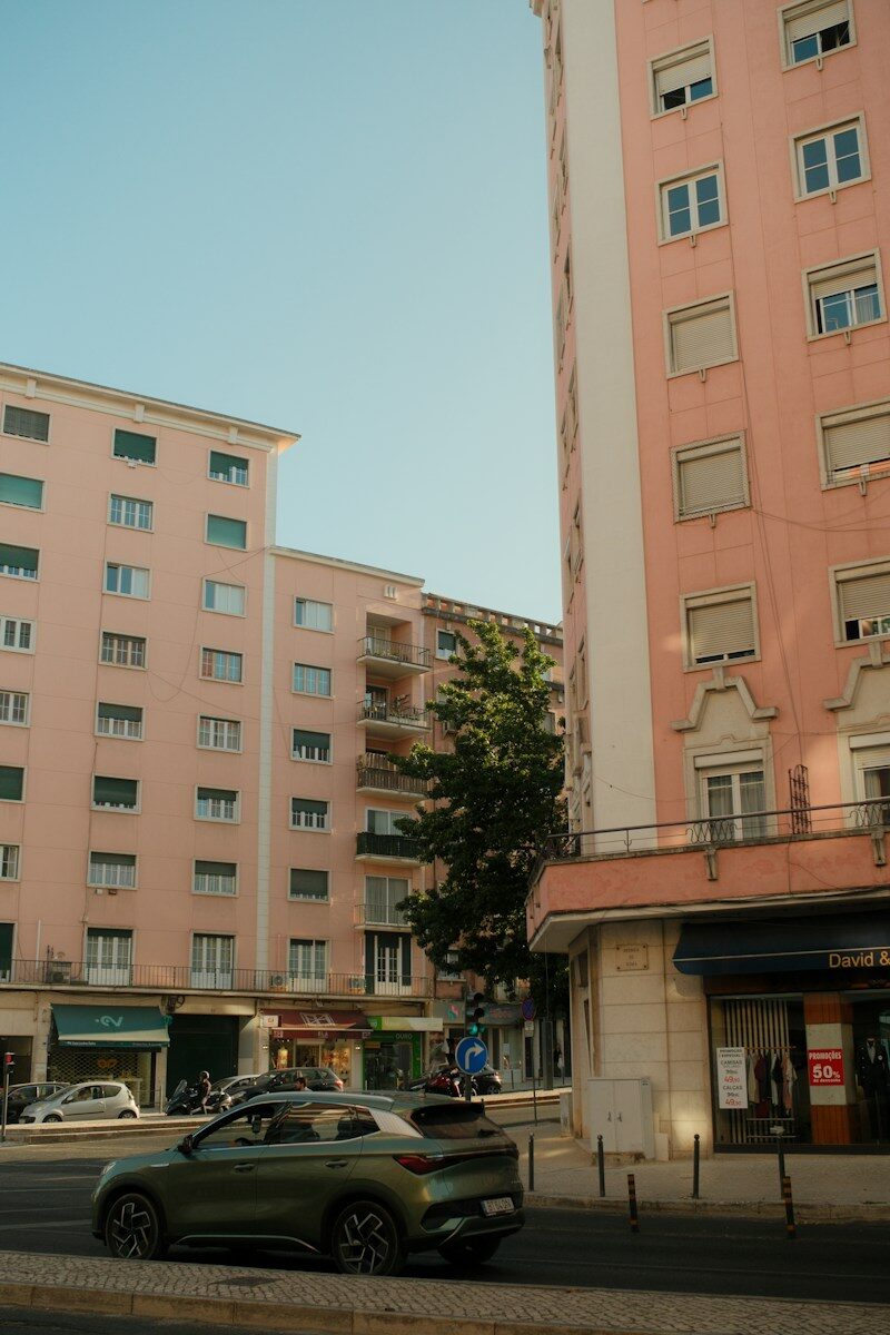 Photo by Timur Seyfelmlyukov A green car driving past pink apartment buildings on a city street